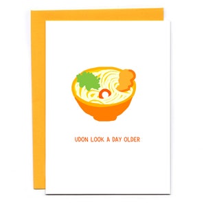 May include: A greeting card with a cartoon illustration of a bowl of udon noodles. The card has the text "Udon look a day older".