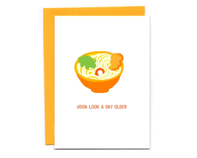 May include: A greeting card with a cartoon illustration of a bowl of udon noodles. The card has the text "Udon look a day older".