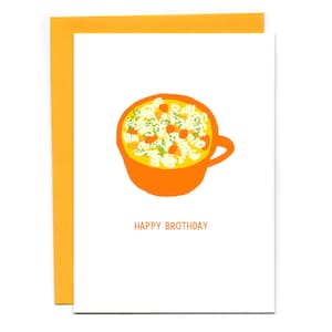 May include: A white card with an orange bowl of yellow noodle soup with green herbs and orange carrots. The text "Happy Brothday" is printed in black below the bowl.