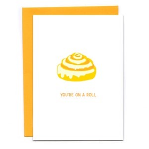 May include: A yellow and white cinnamon roll with the text "You're on a roll" on a white card.