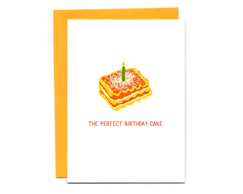 May include: A white greeting card with an orange envelope. The card features a lasagna illustration with a lit green candle. The text "THE PERFECT BIRTHDAY CAKE" is printed in orange below the lasagna image.