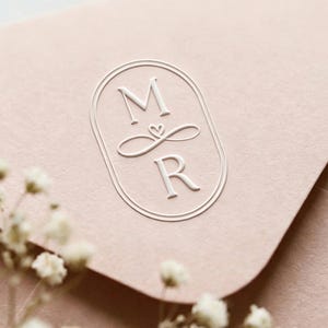 Può includere: Una busta rosa chiaro con un monogramma bianco in rilievo con le lettere "M" e "R" all'interno di una cornice ovale. Un piccolo cuore è tra le lettere. Delicati fiori bianchi in primo piano.
