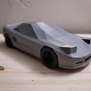 Puede incluir: Un modelo de coche deportivo impreso en 3D de color gris. El coche tiene un perfil bajo y un diseño elegante. El modelo está detallado con líneas y curvas afiladas.