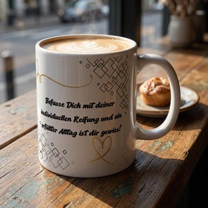 May include: A white ceramic mug filled with coffee featuring latte art. The mug is decorated with black geometric diamond patterns, a gold swirl, a gold heart, and the German text: Befasse Dich mit deiner individuellen Reifung und ein erfüllter Alltag ist dir gewiss!