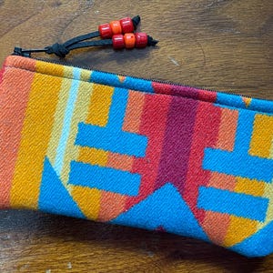 May include: A rectangular, colorful zippered pouch with a geometric pattern in shades of orange, yellow, red, and blue. The pouch has a black zipper and a black cord with orange and red beads attached. The pouch is made of a woven fabric.