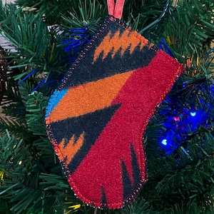 Puede incluir: Un pequeño adorno de calcetín rojo, negro, naranja y azul con un patrón geométrico. El calcetín está colgado de un árbol de Navidad verde.