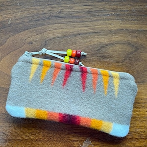 Op de afbeelding: Een kleine, grijze vilten etui met ritssluiting en een kleurrijk geometrisch ontwerp in tinten oranje, geel en rood. De etui heeft een kralen trekker en een witte ritssluiting. De etui is ongeveer 13 cm lang.