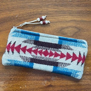 May include: A rectangular, woven pouch with a zipper closure. The pouch features a geometric pattern in shades of red, blue, and brown on a cream background. A white cord with decorative beads is attached to the zipper.