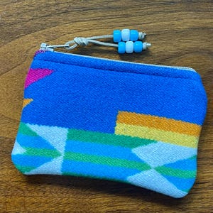 May include: A rectangular zippered pouch featuring a vibrant blue, green, yellow, and white geometric pattern. The zipper pull is a light-colored cord adorned with four small, cylindrical beads in alternating shades of bright blue and white.