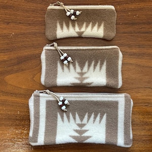 May include: Three rectangular zippered pouches in varying sizes. The pouches are a light brown and cream color with a geometric pattern. Each pouch has a small beaded tassel attached to the zipper pull. The pouches are made of a soft, textured material.