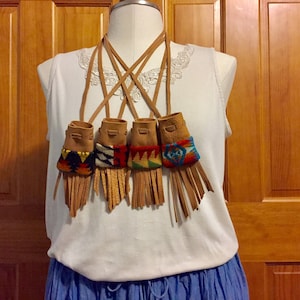 May include: A brown leather necklace with five small pouches. Each pouch is decorated with colorful geometric patterns and has a fringe tassel. The necklace is worn by a person with a white shirt and blue skirt.