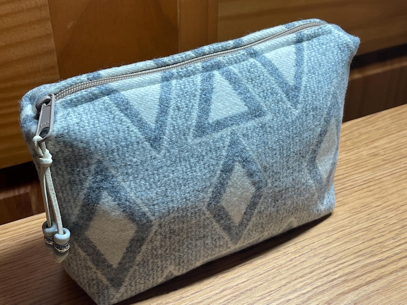 Puede incluir: Un neceser rectangular con un estampado geom&eacute;trico en tonos grises y blancos. La bolsa tiene cierre de cremallera y un peque&ntilde;o lazo con cuentas decorativas. La bolsa est&aacute; hecha de tela texturizada.