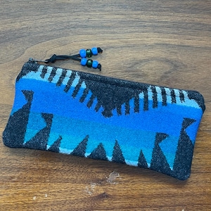 May include: A rectangular zippered pouch featuring a woven pattern of black, bright blue, and teal geometric shapes. The zipper pull is a black cord adorned with two blue and two green plastic beads, providing a functional and decorative accent.
