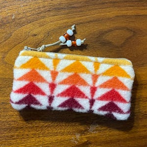 Puede incluir: Un estuche rectangular con cremallera y un patrón geométrico en tonos rojos, naranjas y amarillos sobre fondo blanco. El tirador de la cremallera tiene una cuerda con cuentas blancas y naranjas. El estuche probablemente está hecho de un material suave y texturizado.