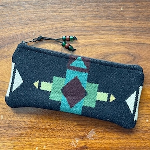 May include: A rectangular black zippered pouch featuring a woven geometric pattern in shades of teal, light green, dark brown, and white. The zipper pull is accented with two small green and brown beads attached to a thin black cord.
