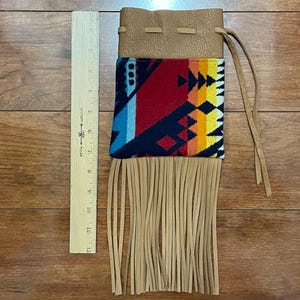 May include: A tan leather pouch with a colorful geometric patterned fabric panel and long fringe. The fabric features red, blue, yellow, and orange designs. The pouch is approximately 12 inches long.