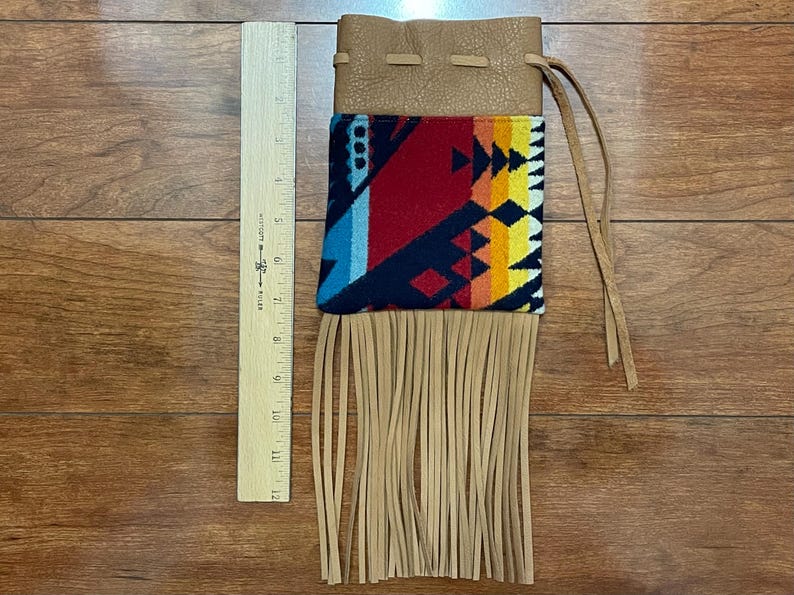 May include: A tan leather pouch with a colorful geometric patterned fabric panel and long fringe. The fabric features red, blue, yellow, and orange designs. The pouch is approximately 12 inches long.