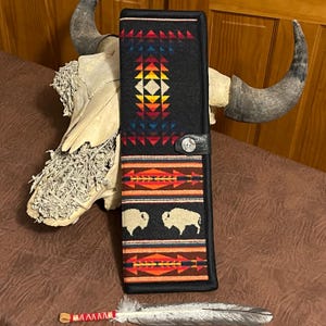 May include: A black case with a Native American design featuring geometric patterns in red, orange, yellow, and blue. The case has a leather strap with a silver button closure and two white bison silhouettes.