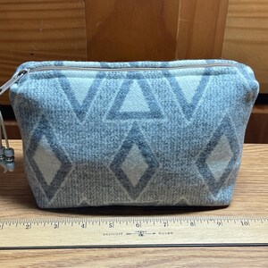 Puede incluir: Peque&ntilde;o neceser rectangular con cremallera, con un patr&oacute;n geom&eacute;trico de diamantes y tri&aacute;ngulos blancos sobre fondo gris. Tiene cremallera plateada y un peque&ntilde;o tirador con cuentas. El neceser mide unos 20 cm de ancho.