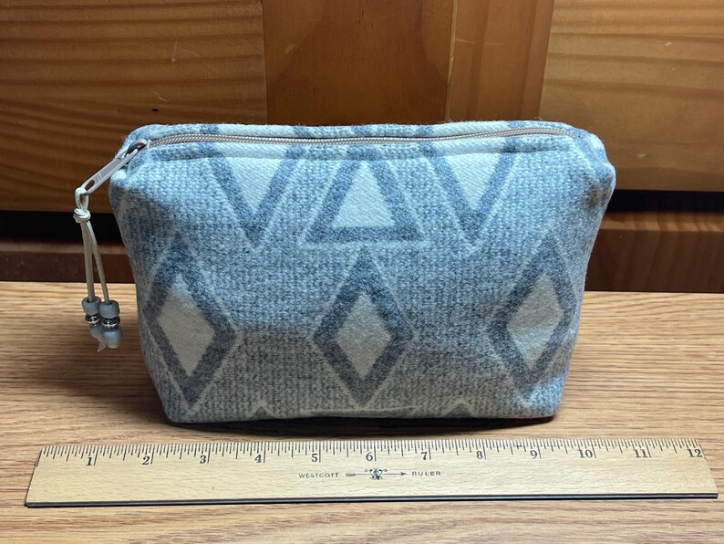 Puede incluir: Peque&ntilde;o neceser rectangular con cremallera, con un patr&oacute;n geom&eacute;trico de diamantes y tri&aacute;ngulos blancos sobre fondo gris. Tiene cremallera plateada y un peque&ntilde;o tirador con cuentas. El neceser mide unos 20 cm de ancho.