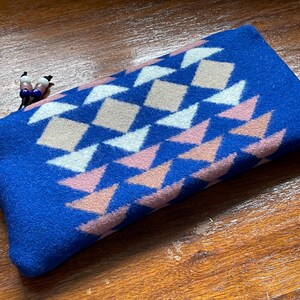 May include: A rectangular blue pouch featuring a geometric pattern of white, pink, and beige diamonds and triangles. The pouch has a zip pull with small decorative beads. The pouch is made of a soft, textured material.