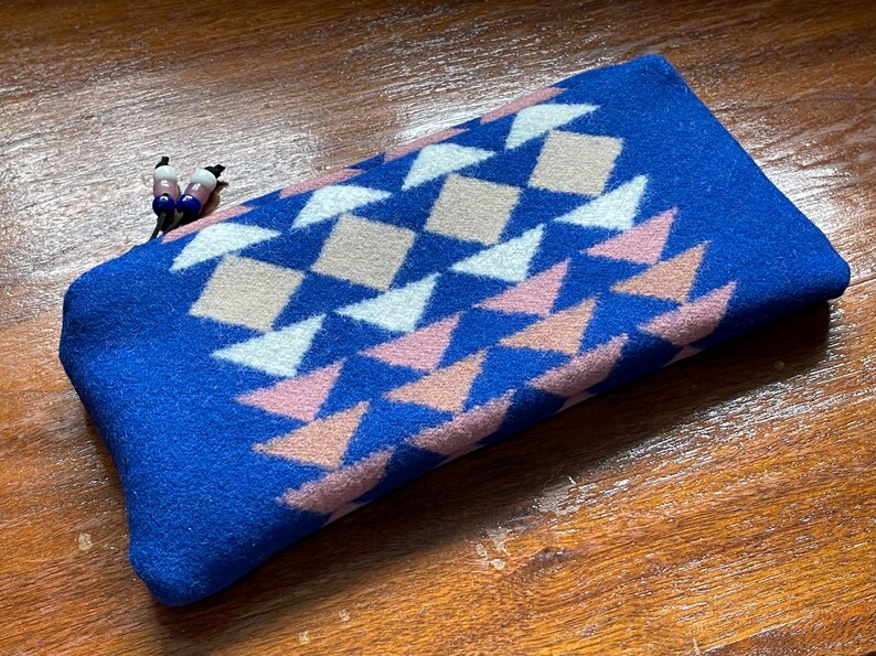 May include: A rectangular blue pouch featuring a geometric pattern of white, pink, and beige diamonds and triangles. The pouch has a zip pull with small decorative beads. The pouch is made of a soft, textured material.