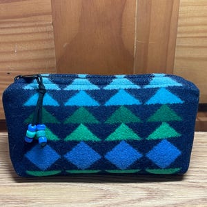 Pode incluir: Uma bolsa com zíper azul marinho com um padrão geométrico de diamantes e triângulos azuis e verdes. A bolsa tem um puxador de zíper preto com contas azuis e verdes. A bolsa é feita de um material macio e texturizado.