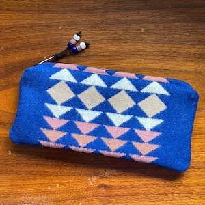 May include: A blue zipped pouch featuring a geometric design in white, pink, and tan. The zip pull has a black cord with decorative beads.