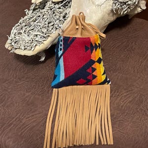 May include: A small, colorful pouch with a geometric pattern in red, blue, and yellow. The pouch has a tan leather drawstring top and a fringe of tan leather strips at the bottom. The pouch is likely used for carrying small items.