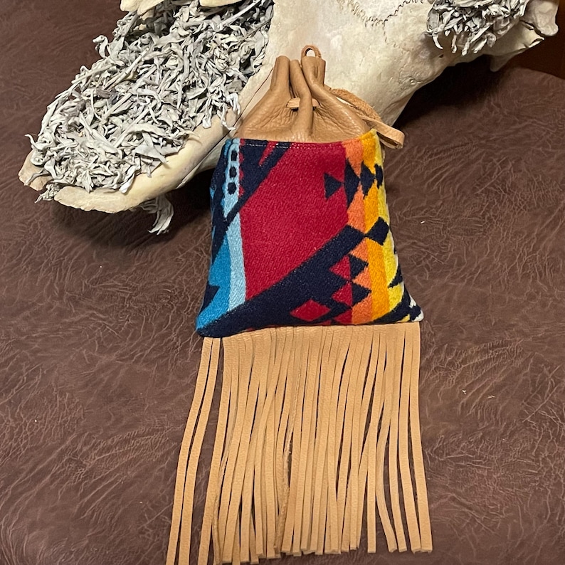 May include: A small, colorful pouch with a geometric pattern in red, blue, and yellow. The pouch has a tan leather drawstring top and a fringe of tan leather strips at the bottom. The pouch is likely used for carrying small items.