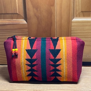 May include: A rectangular, zippered pouch with a colorful, patterned design. The pouch features vertical stripes in shades of red, orange, and yellow, with a central teal stripe. Black arrow-shaped designs are on the front. The zipper pull has decorative beads.