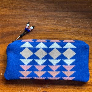 May include: A blue zipped pouch featuring a geometric pattern of white, pink, and tan diamonds and triangles. The pouch has a black zip pull with decorative beads in white, pink, and blue. The pouch is on a wooden surface.