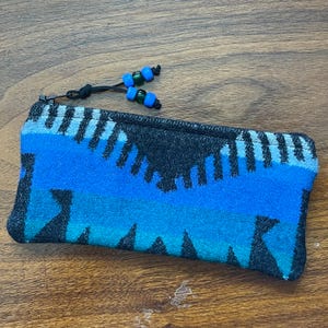 May include: A rectangular zippered pouch featuring a woven pattern in shades of black, royal blue, and turquoise. The zipper pull is a black cord adorned with four small, alternating blue and green plastic beads at the end of the cord.