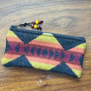 May include: A rectangular zippered pouch featuring a woven geometric pattern in shades of charcoal gray, yellow, orange, and deep red. The zipper pull is accented with two black cords, each finished with a pair of small, round yellow and red beads.
