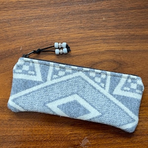 May include: A rectangular pencil case with a geometric pattern in light gray and white. The zipper is black, and the pull has decorative white and silver beads. The case is made of a soft fabric.