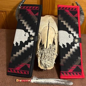 May include: Two black and red patterned cases with a white bison design. The cases have a leather strap closure. A feather and a decorated wooden piece are in the foreground. A skull and horns are in the background.