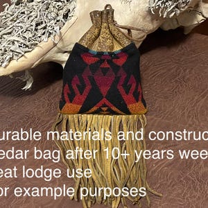 May include: A small, fringed bag with a black, red, and orange geometric design. The bag has a drawstring closure and is made of durable materials. The bag is shown with a light-colored, textured background.