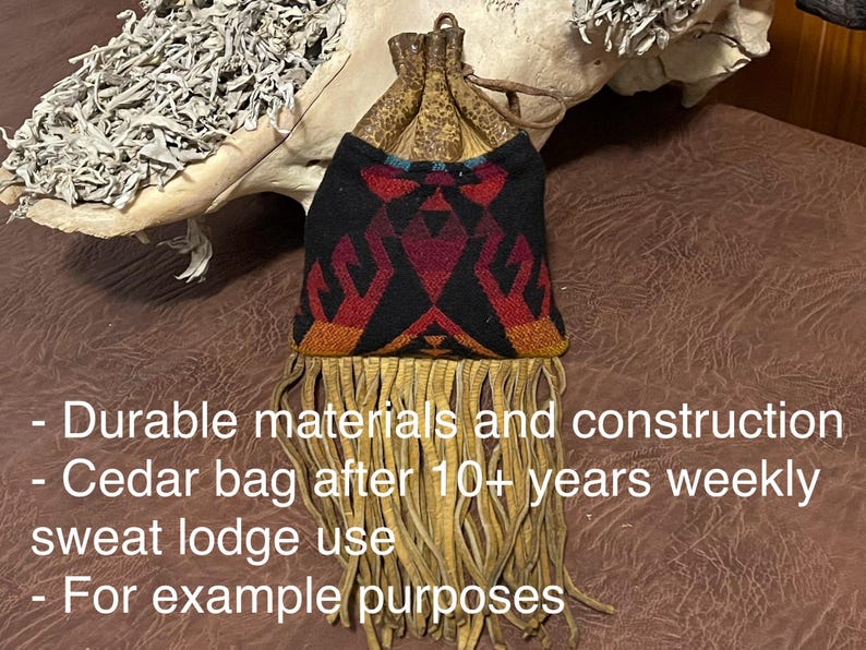May include: A small, fringed bag with a black, red, and orange geometric design. The bag has a drawstring closure and is made of durable materials. The bag is shown with a light-colored, textured background.