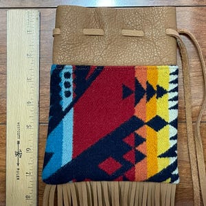 May include: A tan leather pouch with a colorful patterned fabric panel. The fabric features a geometric design in red, blue, yellow, and orange. The pouch has a drawstring closure and a fringe detail at the bottom. A wooden ruler is next to the pouch.