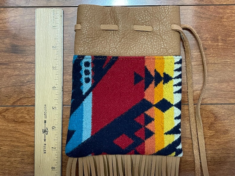 May include: A tan leather pouch with a colorful patterned fabric panel. The fabric features a geometric design in red, blue, yellow, and orange. The pouch has a drawstring closure and a fringe detail at the bottom. A wooden ruler is next to the pouch.