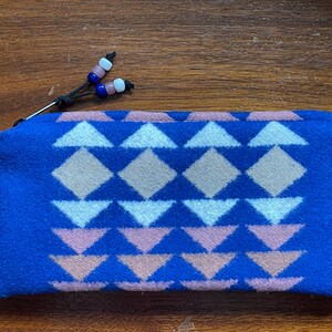 May include: A blue zipped pouch featuring a geometric pattern in white, pink, and tan. The pouch has a black zip pull with pink, blue, and white decorative beads. The pouch is made of a soft, textured material.
