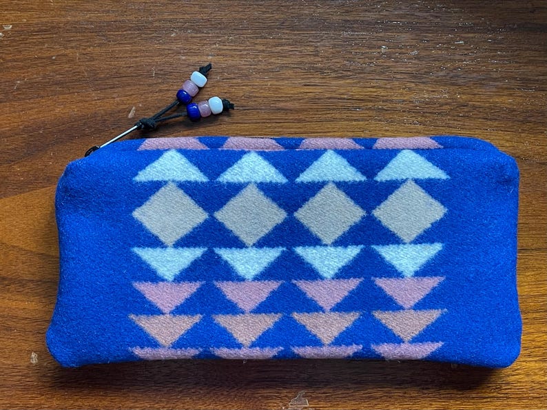 May include: A blue zipped pouch featuring a geometric pattern in white, pink, and tan. The pouch has a black zip pull with pink, blue, and white decorative beads. The pouch is made of a soft, textured material.