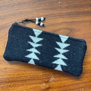 May include: A rectangular zippered pouch made of black felted material featuring two vertical columns of white triangular geometric shapes. The zipper pull is accented with black leather cords and alternating white and black plastic beads.