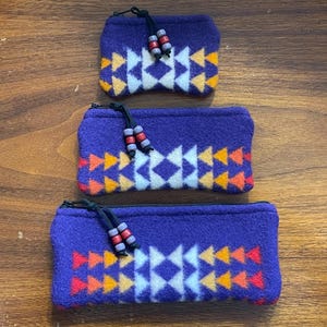 Puede incluir: Tres bolsas de forro polar morado con un patrón geométrico en blanco, amarillo, naranja y rojo. Cada bolsa tiene un tirador de cremallera negro con cuentas decorativas. Las bolsas están dispuestas sobre una superficie de madera.