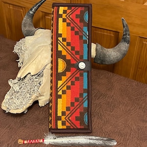 May include: A rectangular, patterned case with a geometric design in shades of red, orange, yellow, and blue. The case has a brown border and a leather strap with a button closure. A decorative feather and a skull with horns are also visible.