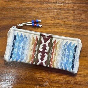 May include: A rectangular pencil case with a colorful geometric pattern in shades of blue, brown, and cream. The case has a zipper closure and a white cord with blue and red beads attached.