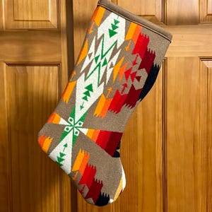 May include: A holiday stocking featuring a tan base color with a vibrant geometric pattern in shades of white, green, orange, red, and dark blue. The design includes repeating arrow and star-like shapes across the fabric surface.