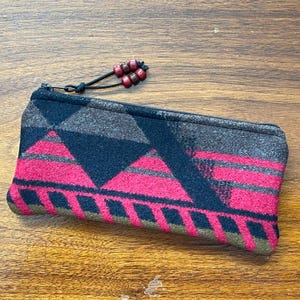 May include: A rectangular zippered pouch featuring a geometric pattern in shades of black, dark gray, and vibrant pink. The zipper pull is a black cord adorned with four small, smooth, dark red beads, providing a functional and decorative accent.