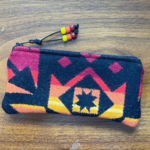 May include: A rectangular, zippered pouch with a Southwestern-inspired geometric pattern in shades of black, red, orange, and yellow. The zipper pull features colorful beads. The pouch is likely used for storing small items.