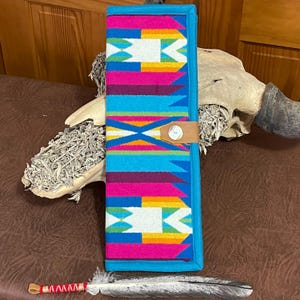 May include: A turquoise blue fabric pouch with a colorful geometric pattern. The pouch has a brown leather strap with a button closure. The pouch is lying on a brown surface with a white feather and a red and white beaded string.